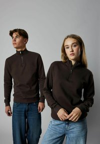 Brązowa bluza na zamek błyskawiczny z długimi rękawami, ściągaczami przy rękawach i u dołu. Dwóch modeli stoi na jednolitym tle, ubrani w casualowe niebieskie dżinsy.