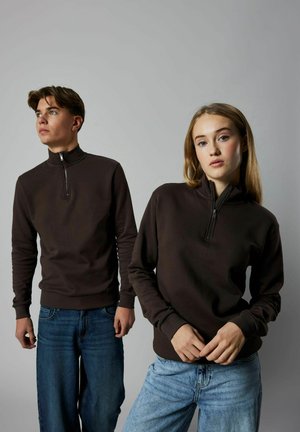 Rjava zip-up majica s dolgimi rokavi, z rebrastimi manšetami in spodnjim robom. Dva modela stojita proti enobarvni ozadju, oblečena v sproščene modre kavbojke.