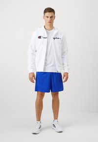 Hvit zip-up hettejakke med marineblå logo, brukt over en hvit t-skjorte og kombinert med blå shorts. Hvite joggesko fullfører antrekket.