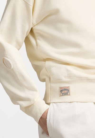 Hanorac de culoare crem, fabricat din material moale, cu manșete cu nervuri, detaliu cu fante pe lateral și o patch personalizată pe marginea inferioară.