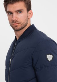 Námořnická modrá vycpaná bomber bunda se zipovým zapínáním, žebrovaným límcem a texturovanou nášivkou na levém rameni se vzorem štítu.