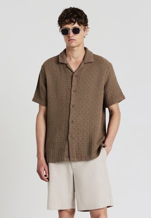 Brown textured short-sleeve shirt with a collared neck, button front, paired with beige pinstriped shorts. Model wears dark sunglasses.