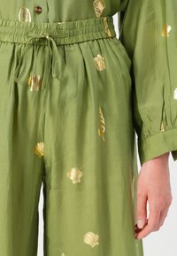 Zelena obleka iz blaga z ohlapnim krojem, elastičnim pasom in zlatimi vzorci školjk. Rokavi so na manšetah nabirani, kar poudari teksturo.