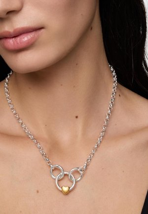 Primo piano del collo con una collana a catena d'argento con tre anelli intrecciati e un piccolo ciondolo a forma di cuore d'oro al centro.