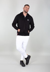 Felpa nera con zip e tasche frontali, abbinata a pantaloni bianchi a sigaretta e sneakers nere. Sfondo grigio chiaro semplice.