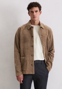 Bruine corduroyjas met een collerug, knoopsluiting aan de voorkant en een zak met een lap aan de linkerkant. G gedragen over een licht shirt en een donkere broek.