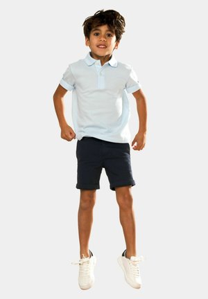 Polo bleu clair avec des manches retroussées, shorts navy et baskets blanches. La chemise présente un col boutonnière et une texture de tissu douce.