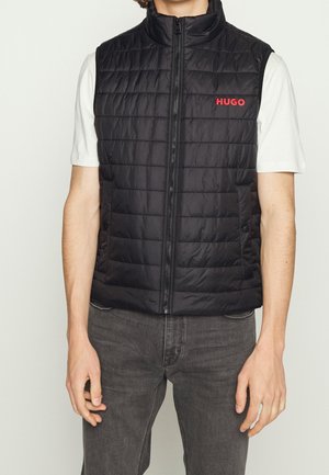 Man wearing black quilted sleeveless vest with red "HUGO" logo over white short-sleeve shirt and dark gray jeans, standing against plain background.