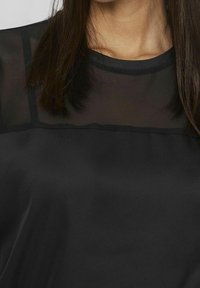 Blouse noire avec un panneau en maille transparente au niveau du col. Texture satinée avec une finition lisse, manches longues, et coupe décontractée.