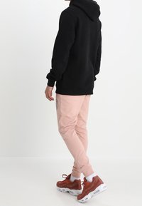 En person som bär en svart huva, ljusrosa sweatpants med sidologga, vita strumpor och rostfärgade sneakers, står mot en vit bakgrund.