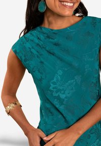 Teal blouse met bloemtextuur en een bootnecklijn. Geaccentueerd met een gouden bloemenarmband en teal oorbellen. Gladde stof, mouwloos ontwerp.