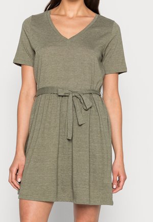 Kobieta ubrana w oliwkowo zieloną sukienkę na krótki rękaw z dekoltem w serek i paskiem w talii, stojąca na tle jasnego, jednolitego tła.