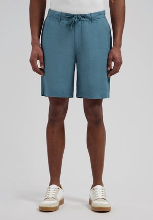 Mannelijk model draagt teal linnen shorts met trekkoord en witte sneakers, staand tegen een effen lichtgrijze achtergrond.