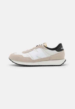 Biało-beżowe buty sportowe New Balance z czarnym kołnierzem, zamszowymi detalami, białym logo "N" i czarną gumową podeszwą, widok z boku na jednolitym tle.