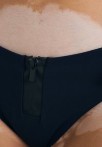 Tamsiai mėlynos spalvos bikini kelnaitės su užtrauktuku priekyje, lygiu paviršiumi ir vidutiniu kirpimu, papuoštos kontrastingomis detalėmis.