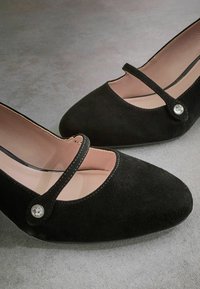 Pantofi Mary Jane din suede negru, cu vârf ascuțit, călcâi închis și o curea pentru gleznă decorată cu un buton ornamental. Captuseala de un roz moale este vizibilă.