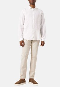 Witte linnen blouse met lange mouwen en knopen aan de voorkant, gecombineerd met beige broek en bruine instappers, staand tegen een neutrale achtergrond.