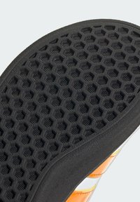 Semelle de chaussure en caoutchouc noir avec un motif hexagonal, comportant des sections transparentes et des accents orange sur le côté. Détails de texture pour une meilleure adhérence visibles.