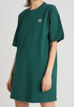 Personne portant une robe T-shirt Adidas ample de couleur vert foncé avec un petit logo Adidas blanc sur la poitrine, sur un fond uni.
