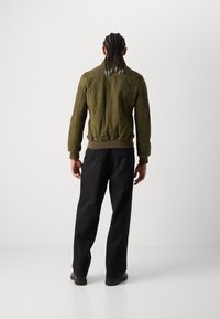 Giacca bomber in suede verde oliva con polsini e orlo a costine, abbinata a pantaloni neri a gamba dritta. Texture liscia con dettagli minimi.
