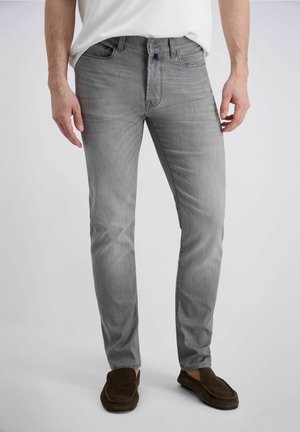 Mann trägt hellgraue Slim-Fit-Jeans mit einem weißen Hemd, das eingesteckt ist, steht und trägt dunkelbraune Slipper.