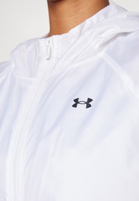 Balta lengva striukė su gobtuvu, turinti užtrauktuką priekyje ir mažą juodą Under Armour logotipą ant krūtinės. Lygus paviršius ir subtilus raštas.
