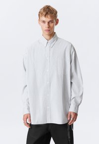 Jovem com cabelo curto loiro, vestindo uma camisa branca com finas riscas verticais cinzas e calças pretas, posando contra um fundo branco.