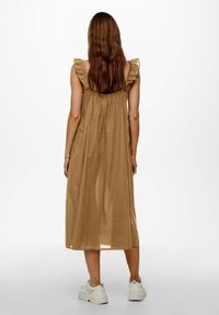 Robe marron mi-longue jusqu'aux genoux avec bretelles à volants, coupe ample et texture douce. Portée avec des baskets blanches. Fond uni.