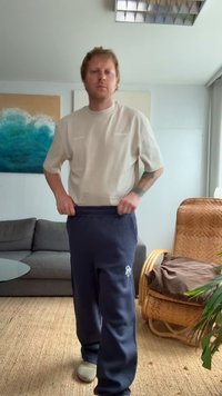 Gai bēšs īsām piedurknēm krekls un tumši zili riboti sporta bikses ar logo. Persona, tērpta pelēkās klogs, stāv telpā ar dīvānu.