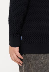 Granatowy sweter z teksturowanej dzianiny z prążkowanymi mankietami i dołem. Posiada małą metkę z logo na dolnej stronie. Noszony na szarych wzorzystych spodniach.