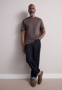 Brown short-sleeve t-shirt paired with dark blue denim jeans. Brown suede sneakers with light gray accents, standing against a light wall.