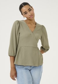Blusa avvolgente verde oliva con tessuto testurizzato, maniche a sbuffo e silhouette a peplo. Abbinata a jeans in denim blu chiaro.