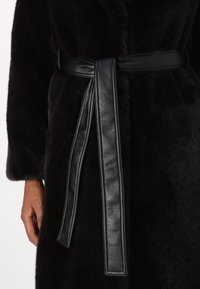 Abrigo de piel negra con una textura suave, que presenta un amplio cinturón de cuero atado a la cintura. El abrigo tiene un acabado esponjoso y mangas ligeramente oversized.