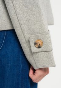 Manica di giacca in lana grigia con chiusura a bottone di colore marrone chiaro. Presenta una texture fine e una cucitura dettagliata lungo la cucitura.