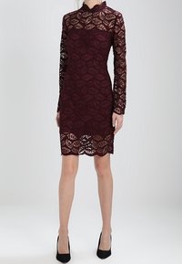 Burgundské krajkové šaty s vysokým výstřihem, dlouhými rukávy a květinovým vzorem; úzký střih s vroubkovaným lemem. Nosit s černými podpatky.