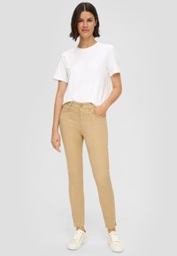 Svetlo bež hlače slim kroja, z stranskimi žepi, gumbnim zapiranjem in poudarjenimi razporki pri gležnjih, v kombinaciji z belo majico.