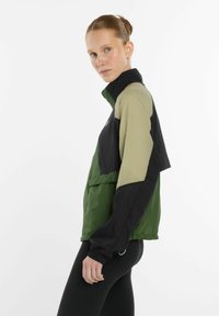 Vrouw met teruggetrokken haar, gekleed in een groen, beige en zwart kleurblokken jas en zwarte leggings, zijdelings staand tegen een witte achtergrond.