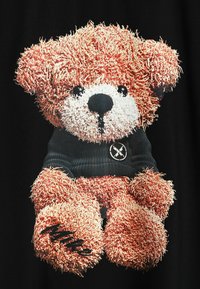 Gráfico de oso de peluche impreso en tela negra; textura de pelaje en marrón rojizo; la ropa negra presenta un emblema circular y un acento manuscrito.