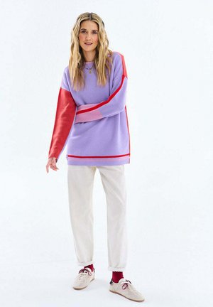 Kobieta o długich blond włosach, ubrana w fioletowo-czerwony sweter, białe spodnie, czerwone skarpetki i białe tenisówki, stojąca na białym tle.