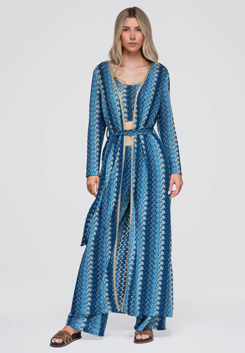 Femme portant un long cardigan bleu à motifs et un crop top assorti avec un pantalon à jambes larges et des sandales marron faciles à enfiler.