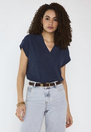Giovane donna con capelli ricci indossa una maglietta blu navy a scollo a V, jeans a vita alta azzurri, cintura marrone e braccialetti e anelli dorati, in piedi su sfondo bianco.