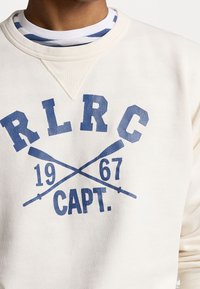 Krämfärgad sweatshirt med en blå grafisk design som har "R.L.R.C 1967 CAPT." och korsade åror. Strukturerad tyg med ribbstickad krage.
