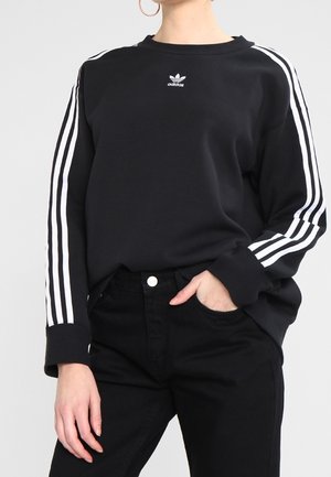 Persoon draagt een zwart Adidas sweatshirt met witte drie strepen op de mouwen en een klein logo op de borst, gecombineerd met een zwarte broek.