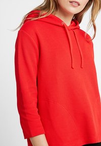 Sudadera roja con tejido texturizado, presentando un bolsillo delantero, una capucha con cordones y mangas de tres cuartos.