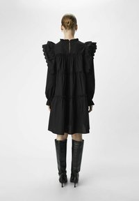 Schwarzes Baumwollkleid mit Rüschensäumen, gestuften Design und einer kleinen Öffnung hinten. Getragen mit kniehohen schwarzen Stiefeln mit Absatz.