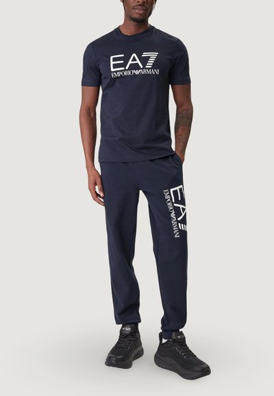 Тъмносиня памучна тениска с бяло лого "EA7 Emporio Armani" на гърдите; комбинирана с тъмносини спортни панталони с лого на бедрото и черни маратонки.