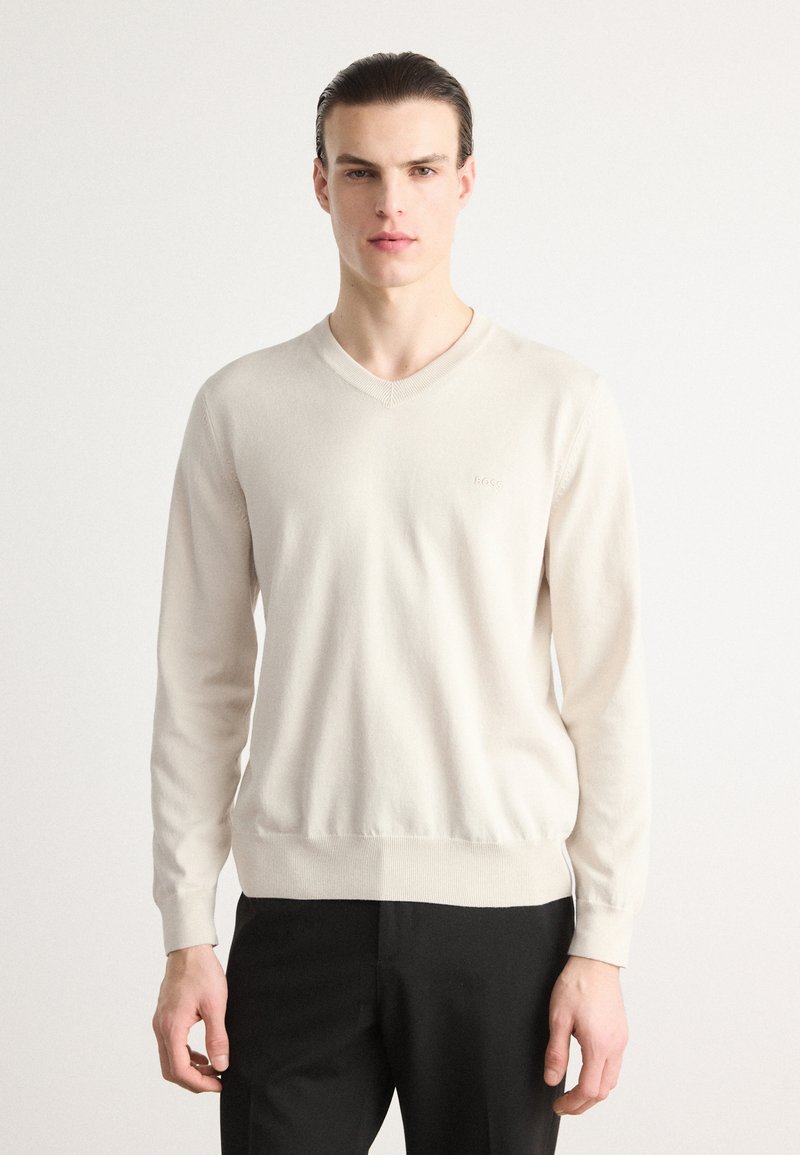 Lys beige V-hals genser laget av glatt, mykt stoff med lange ermer og en tettsittende kant. Diskret logo detalj på brystet.