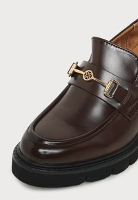 Gros plan d'un mocassin en cuir marron foncé avec une semelle noire épaisse et une barre décorative en métal doré à l'avant.