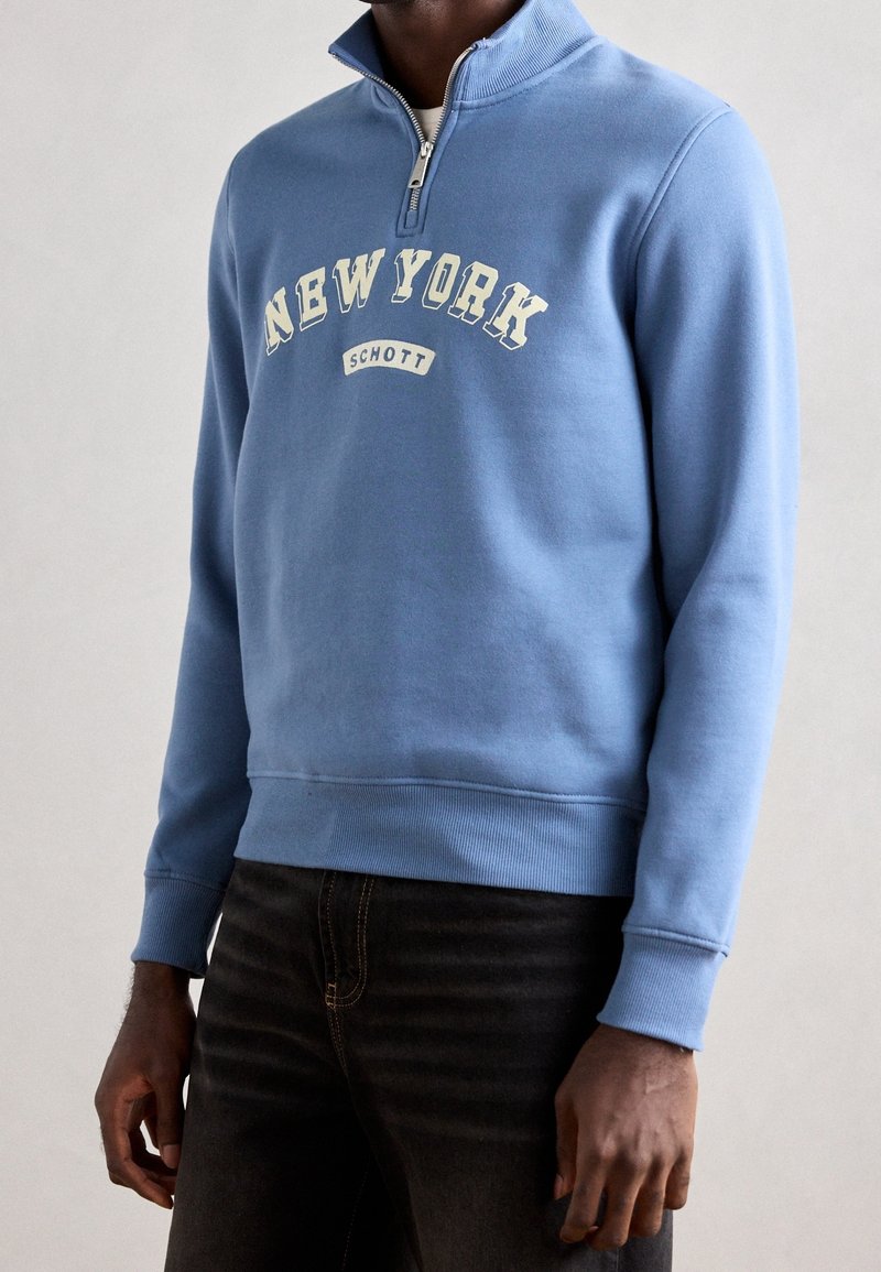 Homme portant un sweat-shirt bleu clair à fermeture éclair quart de zip avec les textes "NEW YORK" et "SCHOTT", associé à un jean foncé, sur fond uni.