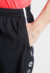 Schwarze Sportshorts mit grauen und weißen Seitestreifen. Sie haben eine Seitentasche und ein sichtbares Tattoo auf der Hand der Person, die sie trägt.